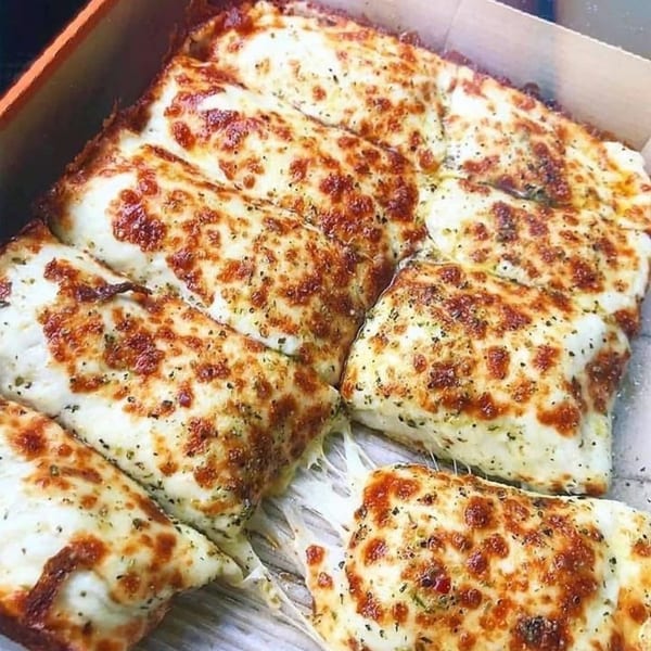Cheesy garlic breadsticks, golden brown and topped with herbs, in a cardboard box.